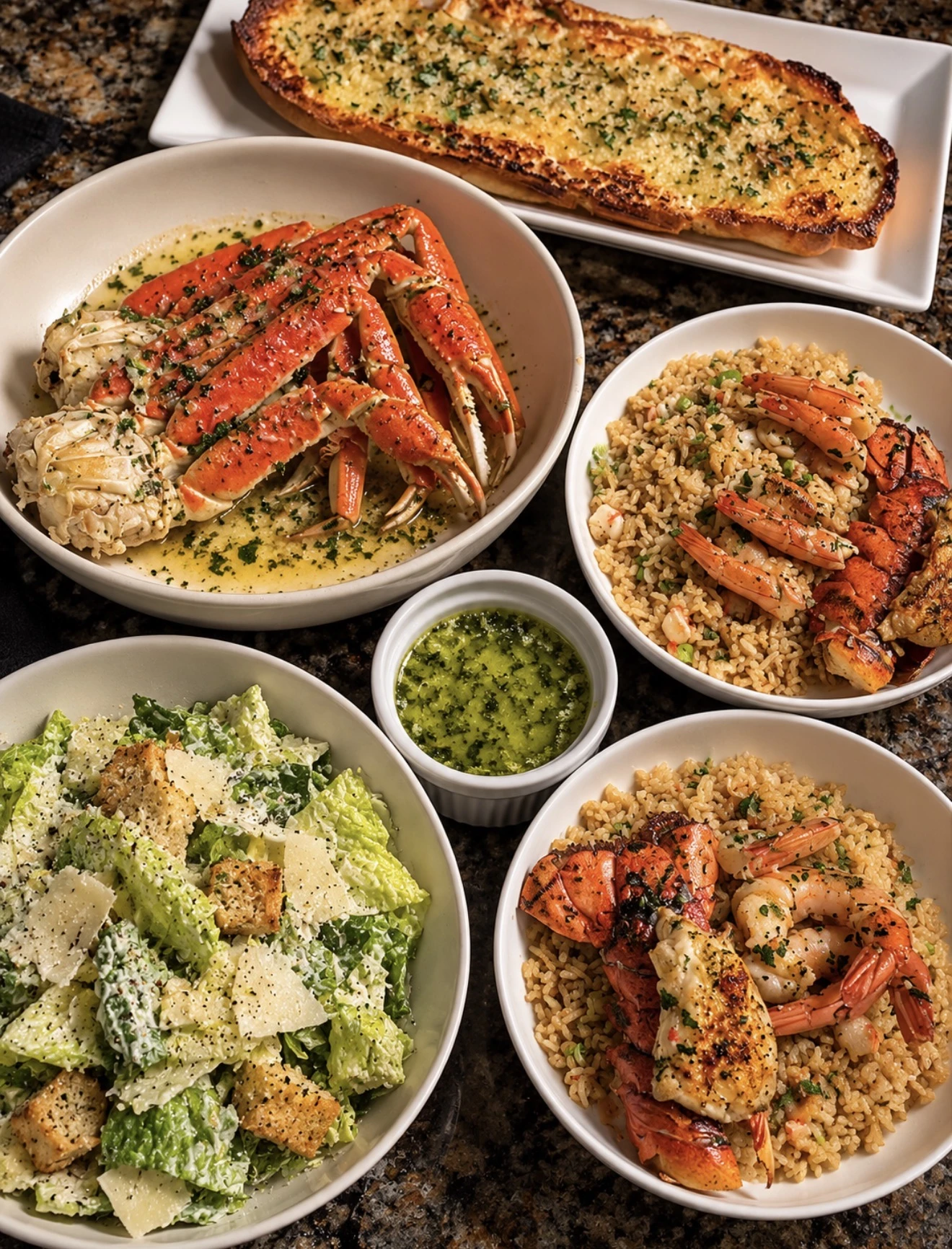 Overhead spread of seafood, pasta, leafy salad, green sauce and a long baked bread topped with herbs.
